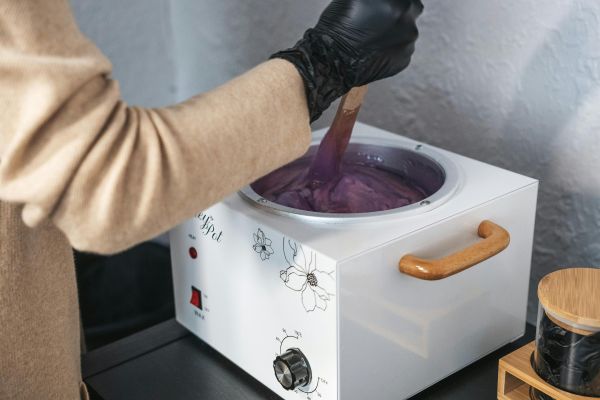 An esthetician wearing gloves stirring purple wax in a wax pot indoors.